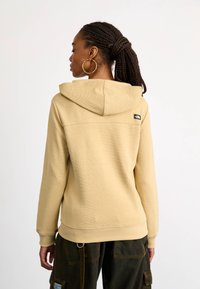 Khaki bluza z kapturem o teksturowanym wykończeniu, z tylnym kapturem, ściągaczami przy mankietach oraz małą naszywką z logo na górnej części pleców. Noszona z ciemnymi spodniami.