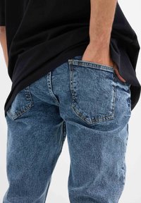 Jeans de mezclilla azul claro con un acabado desgastado, que cuentan con dos bolsillos traseros y costuras visibles, combinados con una camisa negra de corte holgado.