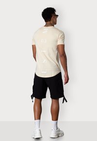 Helles beiges T-Shirt mit kurzen Ärmeln, das senkrechten weißen Text zeigt, kombiniert mit schwarzen Cargo-Shorts. Modell trägt weiße Sneakers und Kniesocken.