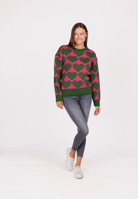 Maglione verde a maglia con un motivo a cuore rosa, colletto rotondo e polsini a coste, abbinato a jeans skinny grigi e sneakers bianche.