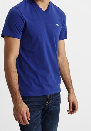 Mężczyzna ubrany w dopasowaną, królewską niebieską koszulkę z dekoltem w serek, z małym zielonym logo krokodyla na lewej piersi, w połączeniu z ciemnoniebieskimi dżinsami.