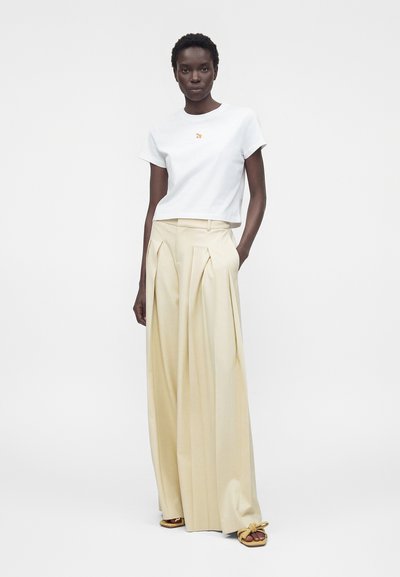 Γυναίκα που φοράει λευκό κοντομάνικο πουκάμισο με μικρή κεντητή λεπτομέρεια, φαρδύ μπεζ πλισέ παντελόνι και μπεζ επίπεδα σανδάλια με φιόγκους.