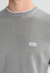 Szary sweter z dzianiny z żeberkowym okrągłym dekoltem, z haftowanym białym logo "BOSS HUGO BOSS" z przodu. Miękka tekstura.