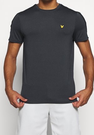 Mężczyzna ubrany w dopasowaną, ciemnoszarą koszulkę z krótkim rękawem z małym żółtym logo ptaszka na lewej piersi, w połączeniu z białymi szortami.