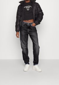 Chaqueta negra acolchada, camiseta negra corta con logo y vaqueros grises. Zapatillas blancas completan el conjunto. Estilo casual y moderno de ropa urbana.