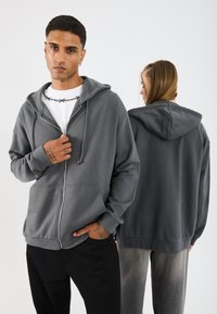 Felpa zip grigia con cappuccio regolabile, tasche frontali e polsini a costine, indossata da due modelli, uno rivolto frontalmente e l'altro girato leggermente di lato.