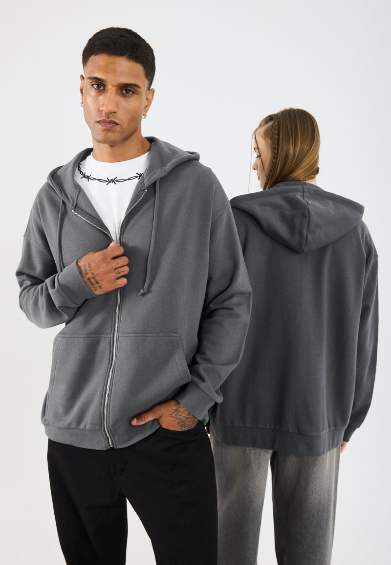 Felpa zip grigia con cappuccio regolabile, tasche frontali e polsini a costine, indossata da due modelli, uno rivolto frontalmente e l'altro girato leggermente di lato.
