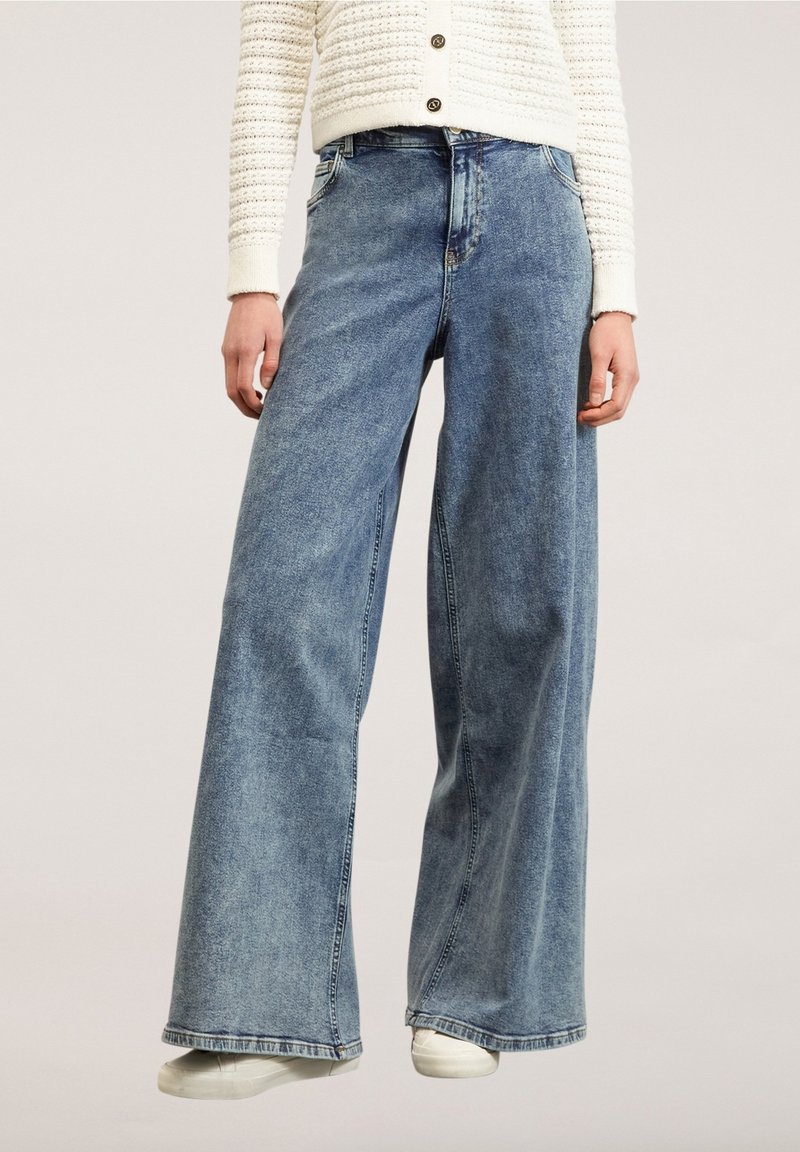 Personne portant un jean large en denim bleu, des baskets blanches et un cardigan crème tricoté avec des boutons.