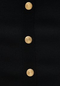 Κοντινή λήψη από μαύρο ριμπ ύφασμα με τρία διακοσμητικά χρυσά κουμπιά που φέρουν σπειροειδές μοτίβο κατά μήκος ενός κάθετου πλακέ.