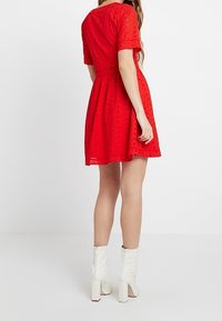 Robe en dentelle rouge avec un corsage ajusté, des manches courtes, une jupe évasée et une taille marquée. Portée avec des bottes blanches à la cheville avec un talon bloc.