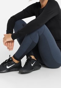 Černý top s dlouhým rukávem, tmavě modré texturované legíny s síťovinovými detaily a černé sportovní boty s bílými akcenty, v sedící pozici.