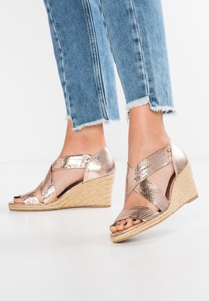 Fødder i lysblå slidte jeans med frynser, iført wedgesandaler i rosaguld med slangeskindslook og flettede hæle af strå på hvid baggrund.