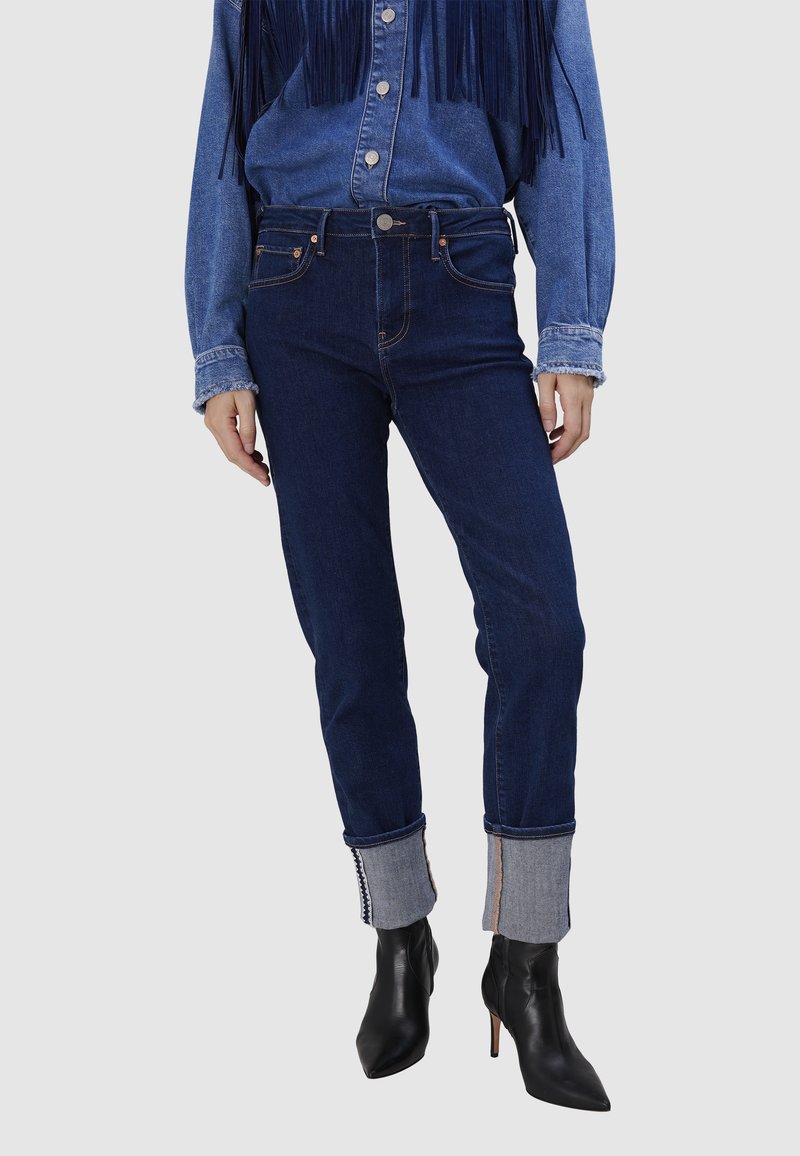 Jeans taille haute bleu foncé avec une silhouette ajustée et un ourlet retroussé. Les retroussages présentent un accent en denim clair avec des détails de couture.