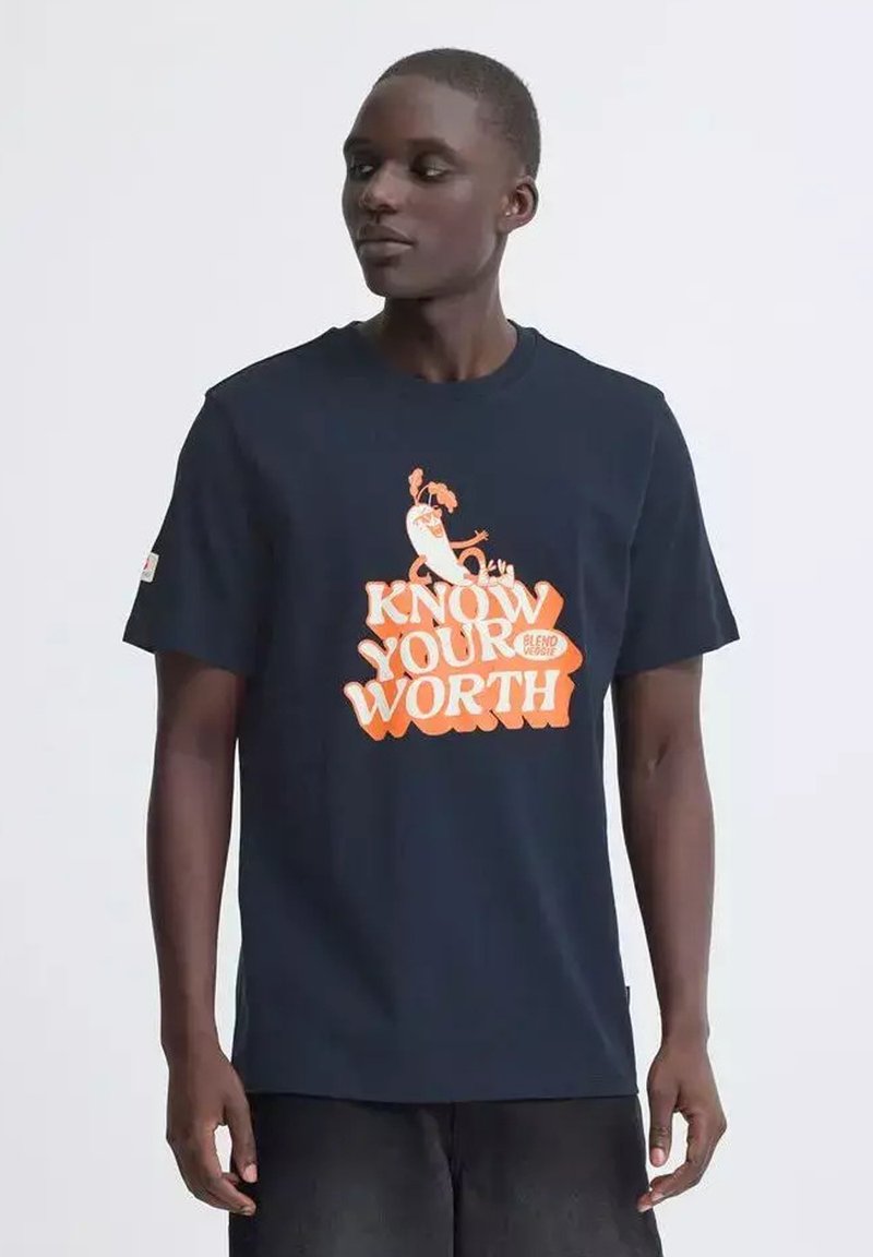 Jeune personne portant un t-shirt foncé avec un motif orange et blanc indiquant "Know Your Worth" et une carotte en dessin animé au-dessus du texte.