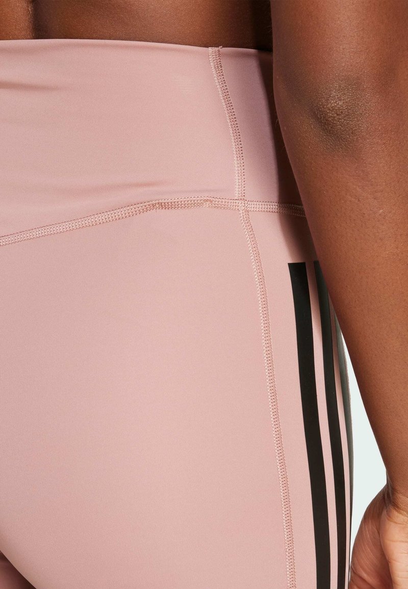 Des leggings taille haute roses présentent des rayures verticales noires sur le côté, une texture lisse et des coutures visibles.