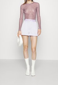 Top de manga larga con un patrón geométrico rosa, combinado con pantalones cortos ajustados color púrpura claro y botas blancas hasta el tobillo. Sosteniendo un bolso blanco.