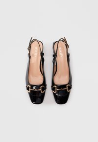 Zwarte gepatineerde leren slingback ballerina's met gouden metalen detail en vierkante neuzen, naast elkaar geplaatst op een lichtgrijze achtergrond.