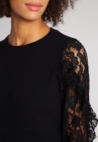 Žena v černém topu s pevným předním dílem a dlouhými květinovými krajkovými rukávy, ukazující částečně tvář a kudrnaté vlasy.