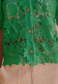 Bluza verde din dantelă brodată, cu modele florale, nasturi aurii și textură transparentă, alături de un strat de bază în culori neutre.