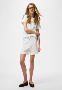 Fiatal nő hosszú hajjal, sárga árnyalatú szemüveget visel, fehér rövid ujjú felsőt, hozzá illő rövidnadrágot és fekete bebújós cipőt, egyik karját keresztbe téve áll.