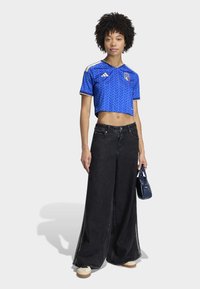 Blue cropped jersey with chevron pattern and Italy crest, paired with wide-leg black jeans and white sneakers, holding a small blue handbag.