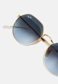Gafas de sol con marco de metal dorado y lentes grandes y redondos de color azul degradado. Puntas de patilla de plástico transparente y logotipo de la marca destacado en la lente.