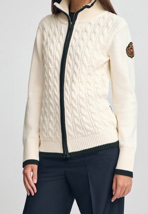 Maglione bianco con cerniera a coste e finiture scure su polsini, orlo e cerniera, indossato con pantaloni scuri, presenta una toppa sulla manica sinistra superiore.