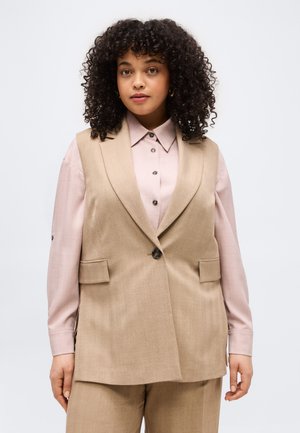 Kobieta o kręconych włosach, ubrana w beżową, bezrękawnikową marynarkę na jasnoróżowej koszuli zapinanej na guziki oraz pasujące beżowe spodnie.