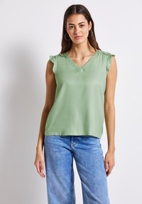 Helles grünes ärmelloses Top mit V-Ausschnitt und Rüschenakzenten an den Schultern, aus glattem Stoff gefertigt, kombiniert mit blauen weiten Jeans.