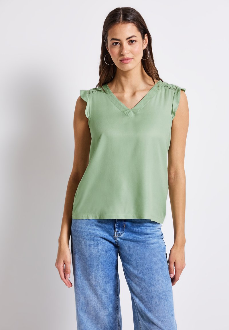 Helles grünes ärmelloses Top mit V-Ausschnitt und Rüschenakzenten an den Schultern, aus glattem Stoff gefertigt, kombiniert mit blauen weiten Jeans.