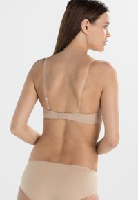 Nude bh med tynne justerbare stropper og en krok-og-øye-lukking, med en glatt tekstur og minimalistisk design.