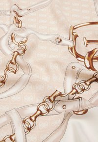 Tejido de seda con fondo beige que presenta un estampado de correas de cuero blancas y eslabones de cadena dorados, suavemente drapeado con leves pliegues.
