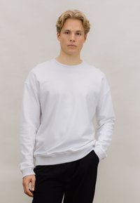 Hvit hettegenser med rund hals og ribbede mansjetter og kant. Enkelt design, laget av mykt stoff, vist med svarte bukser.