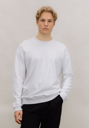 Hvit hettegenser med rund hals og ribbede mansjetter og kant. Enkelt design, laget av mykt stoff, vist med svarte bukser.