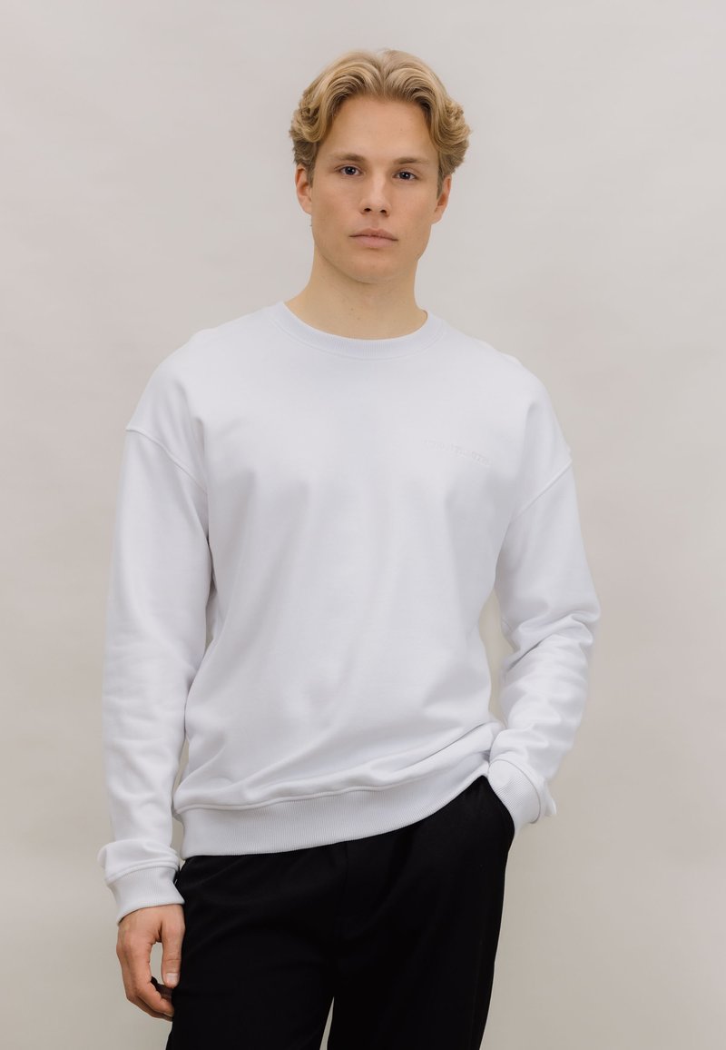 Hvit hettegenser med rund hals og ribbede mansjetter og kant. Enkelt design, laget av mykt stoff, vist med svarte bukser.