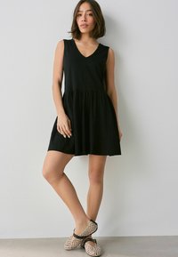 Sort ærmeløs kjole med V-udskæring og samlet talje. Blødt stof med en udvidet nederdel. Båret med mønstrede, lyse flade sko.