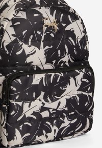 Sac à dos noir et beige avec une finition matelassée, arborant un motif de feuille tropicale et une poche zippée à l'avant. Accent du logo doré visible.