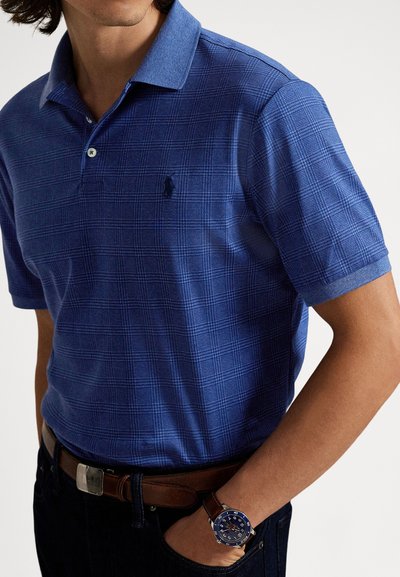 Muž oblečený v modrom károvanom krátkym rukávom polokošeli, hnedom opasku, tmavých džínsach a modrými hodinkami s hnedom koženým remienkom.