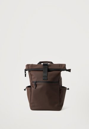 Brauner Rucksack mit oberer Klappe, die durch einen schwarzen Riemen gesichert ist, vordere Reißverschlusstasche und zwei Seitentaschen vor einem einfarbigen hellen Hintergrund.