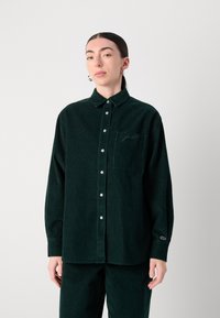 Camicia in corduroy verde con colletto abbottonato, tasca anteriore e logo ricamato sulla manica. Maniche lunghe e tessuto texture.