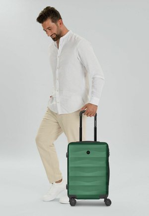 Hombre con camisa blanca y pantalones beige que jala una maleta verde con ruedas, sonriente y mirando hacia abajo, sobre un fondo claro y simple.