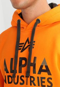 Felpa arancione in cotone con cappuccio regolabile, caratterizzata da scritte grafiche nere "ALPHA INDUSTRIES" e un logo al centro.