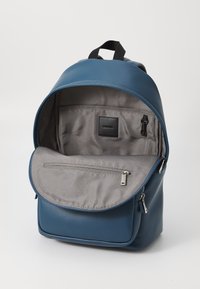 Mochila de cuero azul con un interior de tela gris, bolsillo con cremallera y cierre de solapa; cuenta con un asa superior y correas ajustables.