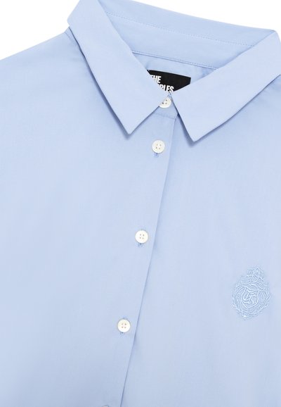 Jasnoniebieska, krótko rękawowa koszula z kołnierzykiem w kształcie spiczastym, z białymi guzikami i haftowanym emblematem na piersi. Gładki, bawełniany materiał.