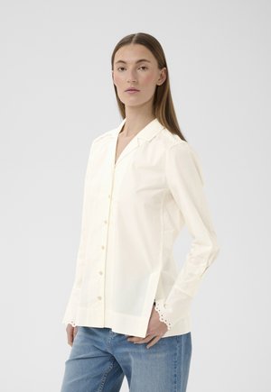 Donna con capelli castani lisci indossa una camicetta crema a maniche lunghe con bottoni, con dettagli a occhiello su polsini e spalle, abbinata a jeans blu.