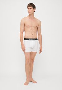Fehér boxer alsónadrág fekete rugalmas derékrésszel, amelyen a "VERSACE" logo látható. Sima pamut anyag, testhezálló kivitel, térd alá érő hosszúság.