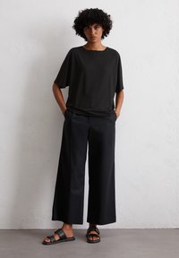 Czarny, luźny top z szerokimi rękawami, zestawiony z ciemnymi spodniami o szerokich nogawkach i czarnymi klapkami na tle jednolitym.