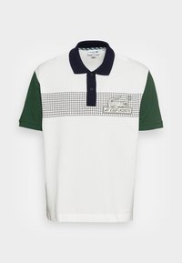 Vit polotröja med korta gröna ärmar. Har en marinblå krage, rutmönster över bröstet och broderad "Le Club Lacoste" logotyp.