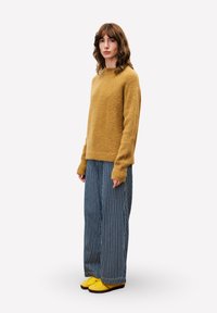 Vrouw met golvend bruin haar die een mosterdkleurige trui, brede blauw-wit gestreepte broek en gele schoenen draagt, zijwaarts staand op een witte achtergrond.