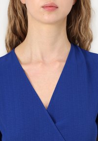 Plava bluza s V-izrezom, teksturiranom tkaninom i kratkim rukavima koji ističu glatku liniju vrata i definirane šavove.